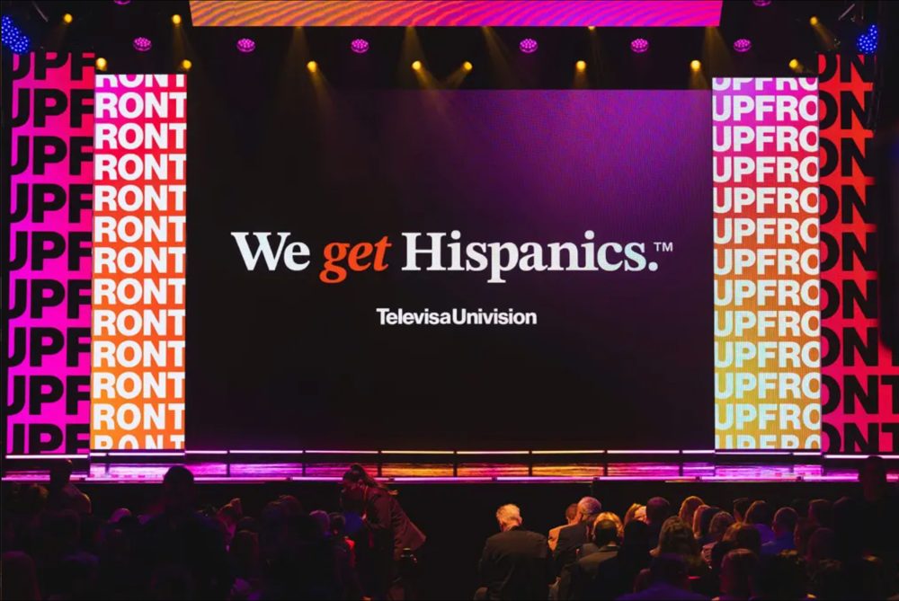 TelevisaUnivision Upfront stage with the line We get Hispanics on the main screen, framed by gradient UPFRONT panels, with the audience in the foreground.