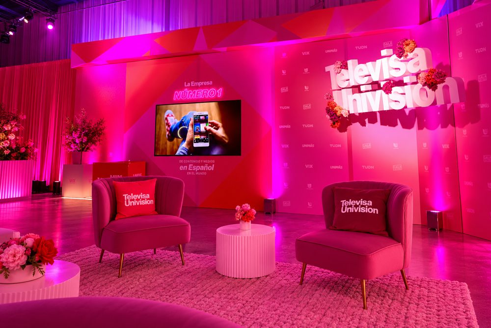 TelevisaUnivision event lounge in pink light: two velvet armchairs with branded pillows, a screen reading La Empresa Número 1, and a 3D TelevisaUnivision logo dressed in flowers.