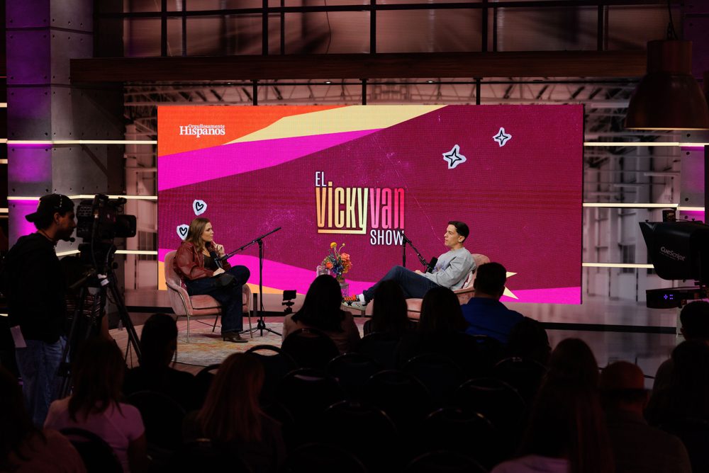 Taping of El Vicky Van Show for Orgullosamente Hispanos: two hosts in armchairs on a colorful stage in front of a live audience, with a camera operator at left.